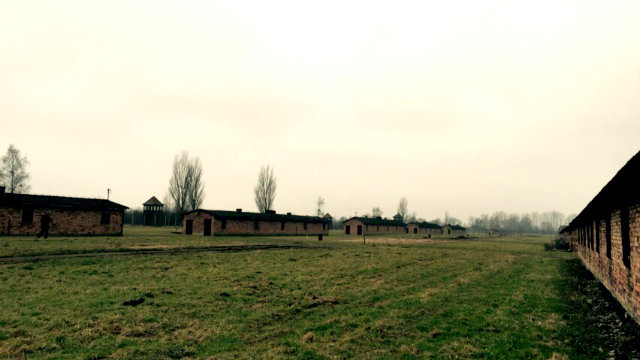 Foto del campo di concentramento di Aushwitz: gli austeri edifici bassi e lunghi in mattoncini destinati ai detenuti si susseguono ordinatamente su un vasto prato. In lontananza le torrette di controllo.